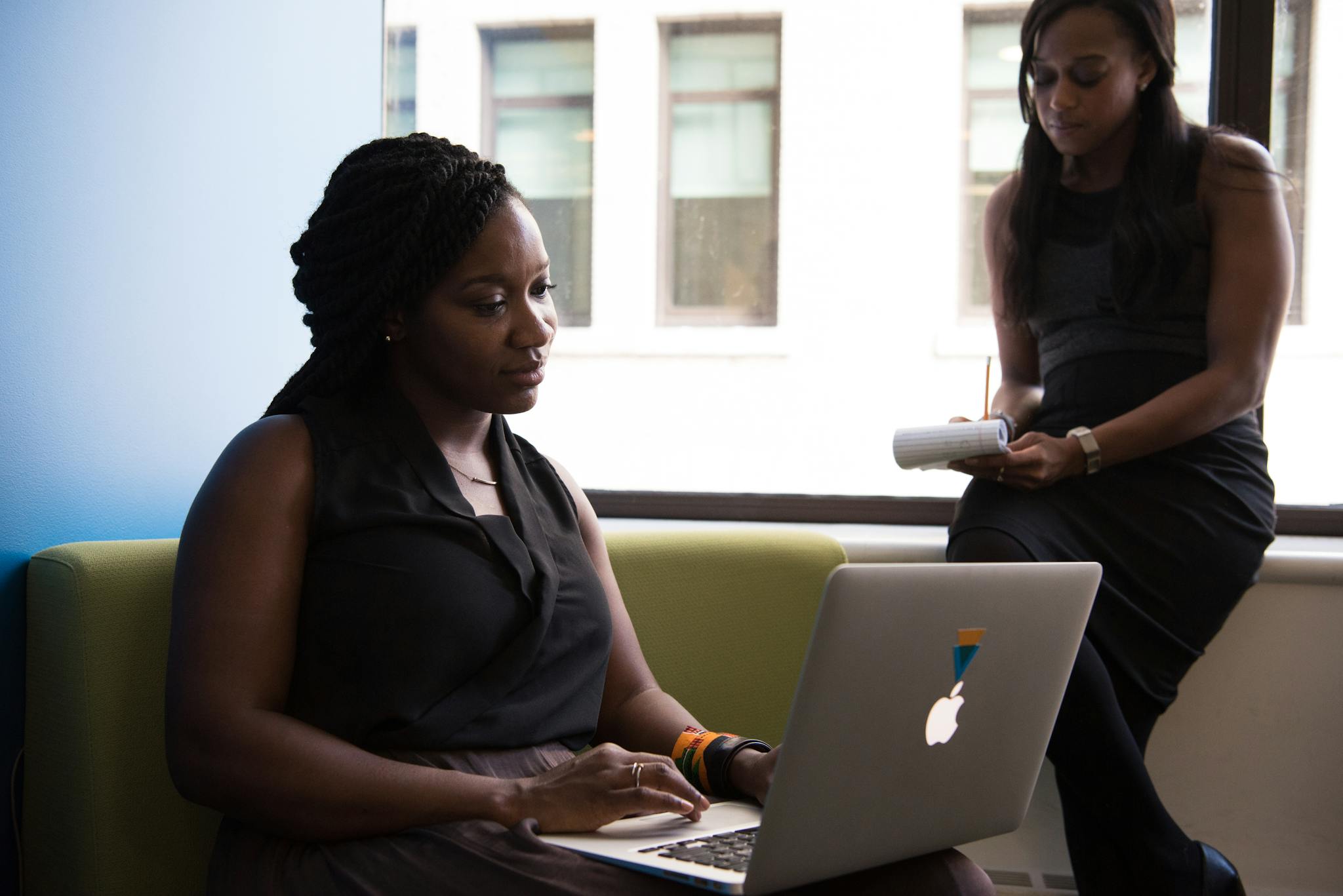 Two women in a business setting working on a laptop and taking notes, fostering teamwork.